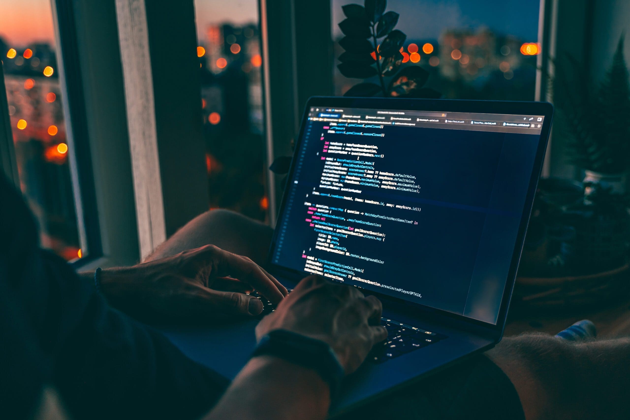 Ein Webentwickler arbeitet bei künstlichem Licht vor einem großen Fenster mit Blick auf eine nächtliche Stadtlandschaft. Auf dem Laptopbildschirm ist Code zu sehen, der auf Web development hindeutet.