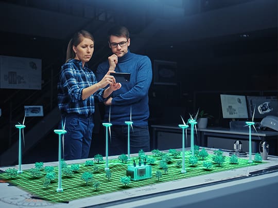 Zwei Ingenieure stehen in einem dunklen Raum und interagieren mit einer 3D-Darstellung einer Windfarm, die durch Extended Reality (XR) Technologie visualisiert wird. Sie betrachten und diskutieren die Daten auf einem Tablet, das die Information in Echtzeit mit der holographischen Darstellung synchronisiert.