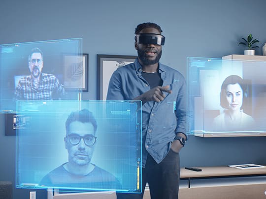 Ein Mann in einem modernen Büro trägt ein XR-Headset und interagiert mit virtuellen 3D-Bildschirmen, die Kollegen in Echtzeit anzeigen, was die Möglichkeiten von Extended Reality für die Telearbeit und Kollaboration demonstriert.