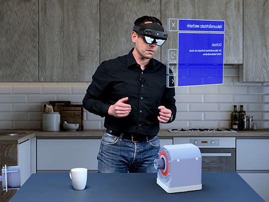 Ein Mann in einem schwarzen Hemd verwendet Extended Reality (XR) Brillen in einer Küche, um ein interaktives, holographisches Display zu bedienen, das über einer elektronischen Vorrichtung schwebt, wobei virtuelle Informationen über dem Gerät angezeigt werden.