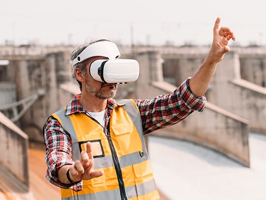 Ein Bauarbeiter in Sicherheitskleidung und mit Helm trägt eine XR-Brille und interagiert mit einer virtuellen Schnittstelle auf einer Baustelle, wobei er Gesten zur Steuerung der Extended Reality-Umgebung verwendet.