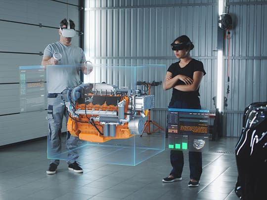 Zwei Personen in einem industriellen Setting, eine davon mit XR-Headset und -Controllern, interagieren mit einer virtuellen Maschinenkomponente, die als Teil eines Extended Reality-Ausbildungsprogramms dargestellt wird.